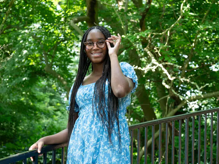 A person with long braided hair and wearing a blue floral dress stands outdoors by a railing, adjusting their glasses and smiling.