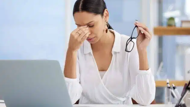 A person in a white shirt is sitting at a desk, holding glasses and rubbing the bridge of their nose, appearing stressed or tired.